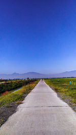 Empty road amidst field against clear blue sky