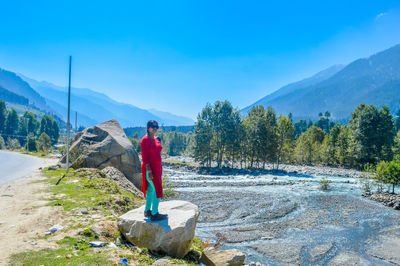 Woman standing on rock at riverbank against blue sky