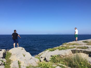 Men standing on rock at beach against clear blue sky