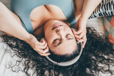 Curly haired plus size young woman listens to music using wireless headphones on bed.