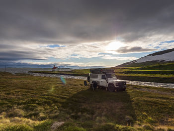 Off-road car parked in front of icelandic river at cloudy sunset with secluded unadsdalskirkja church in background