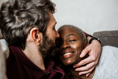 Young bearded man in casual shirt lying on comfortable couch in embrace with african american woman in cozy living room with closed eyes