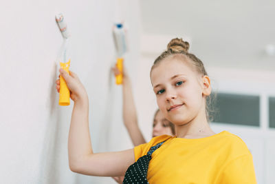 A girl with her sister in bright blue and yellow clothes helps to paint the walls in her room white.