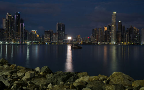 Illuminated buildings by sea against sky at night