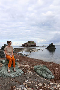 Full length of woman sitting on rock at sea shore against sky