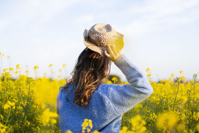 A girl from behind putting on a hat with yellow flowers on field against sky