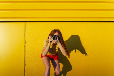 Woman photographing through camera while standing against yellow wall