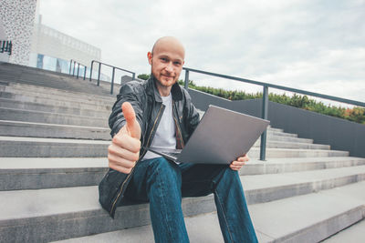 Portrait of smiling businessman with laptop showing thumbs up while sitting on staircase