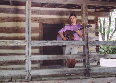 Portrait of handsome man holding acoustic guitar while standing outside log cabin