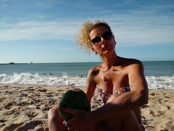 Woman wearing sunglasses while sitting with coconut on sand at beach against sky