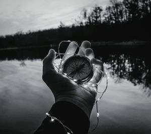 Cropped hand holding compass with illuminated string lights by lake