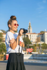 Woman photographing with camera against sky in city