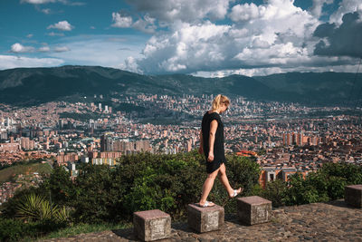 Side view of woman walking stone seats with buildings in backgrounds against cloudy sky