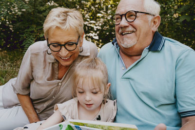 Happy grandparents sitting with granddaughter reading book at park