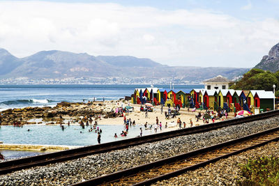 Railroad tracks with people in background at beach against cloudy sky