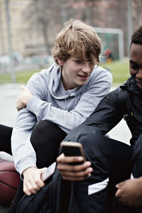 Teenage boy looking at friend's mobile phone while sitting on sidewalk after basketball practice