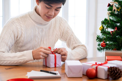 Young man tying ribbon on gifts over table at home