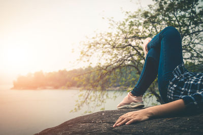 Low section of man sitting by lake against sky during sunset