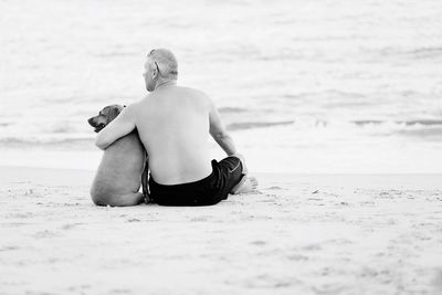 Rear view of shirtless man with dog sitting on beach