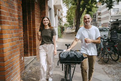 Happy friends wheeling with bicycle while holding disposable cup on sidewalk