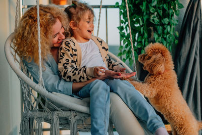 Happy mother and daughter play at home on the balcony, sitting on a hammock chair, with a pet