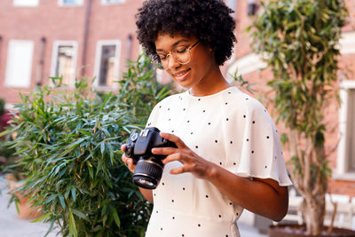 Smiling teenage girl holding camera while standing against plants