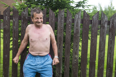 Shirtless senior man standing against fence at yard