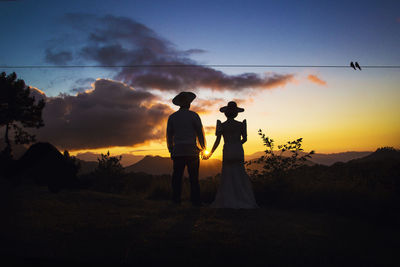 Silhouette couple holding hands while standing against sky during sunset