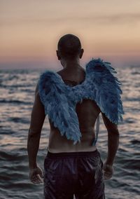 Rear view of shirtless man with costume wings in sea against sky