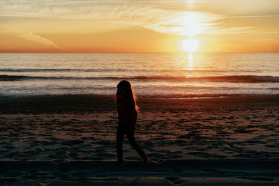 Rear view of woman standing at beach against sky during sunset