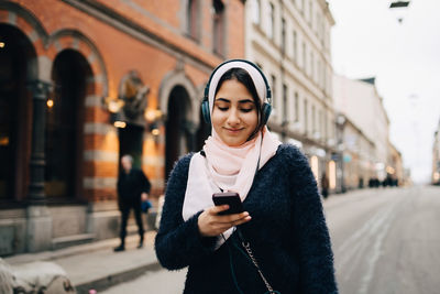 Smiling teenage girl listening to headphones using smart phone while walking on street in city