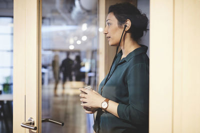 Side view of businesswoman holding smart phone seen from glass at office