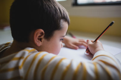 Side view of crop cute kid sitting at wooden table and drawing with pencil on paper while relaxing at weekend at home