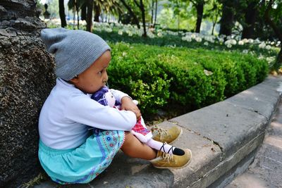 Side view of cute baby girl sitting on retaining wall by plants at park