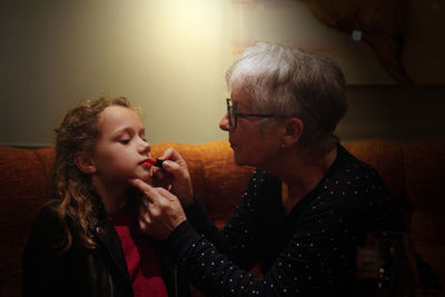 Grandmother applying lipstick to granddaughter while sitting on sofa