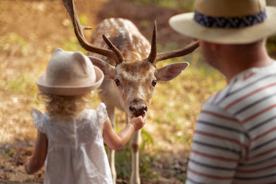 Cute baby girl, toddler, child and man feeding big brown deer, fawn with antlers in forest,park,farm