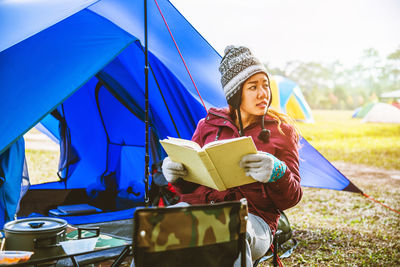Young woman reading book while sitting on chair at campsite
