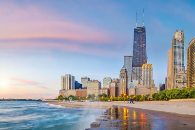 Modern buildings by sea against sky during sunset