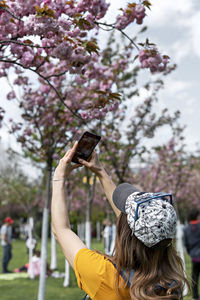 Rear view of  young woman in cap taking photo of blooming sakura almond tree on smartphone in park