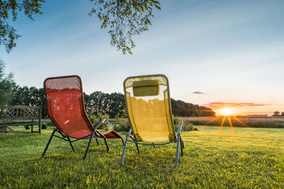 Abandoned chairs on field against sky at sunset