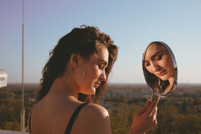 Rear view of young woman with mirror standing against clear sky