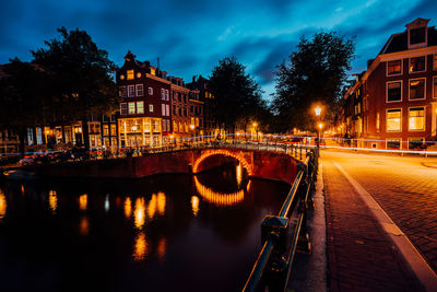 Illuminated bridge over canal by buildings against sky at dusk