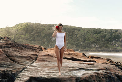 Seductive young woman wearing white swimsuit while standing on rock at beach