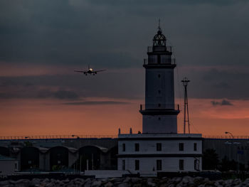 Airplane flying over building against sky during sunset