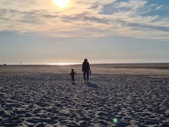 Child and adult on beach against sky during sunset