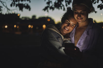 Smiling woman embracing non-binary friend using mobile phone at night