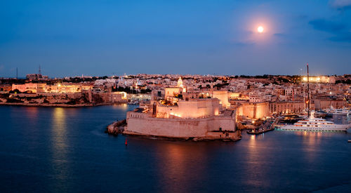 Sea by illuminated buildings against sky at dusk