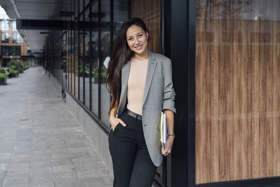 Smiling businesswoman with hand in pocket standing outside office building