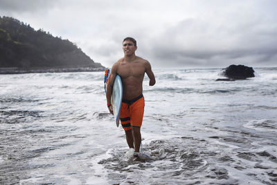 Disabled man carrying surfboard while walking out of sea against sky