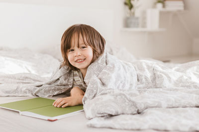 A cute toddler boy is lying on the bed with a book wrapped in a blanket, smiling 